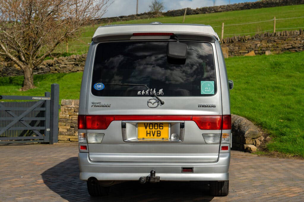 A silver 2006 Mazda Bongo Friendee with a UK registration plate "VO06 HVB" is parked on a brick driveway. The rear window features a family cycling graphic sticker and a circular green sticker. In the background, there's a stone wall and grassy area.