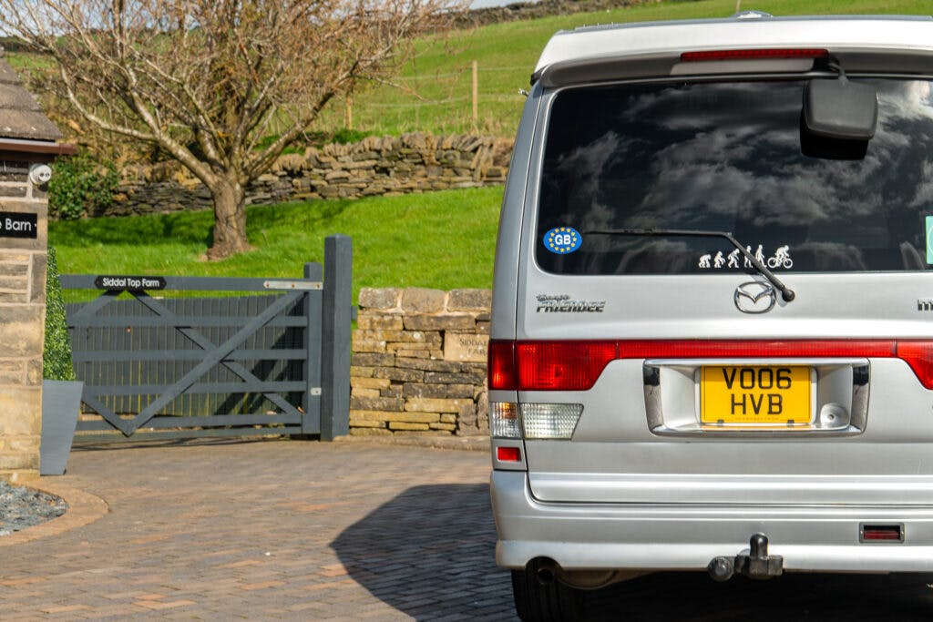 A silver 2006 Mazda Bongo Friendee campervan with the license plate "VO06 HVB" is parked in front of a stone wall and a house entrance gate labeled "Sidda Top Farm." The rear window of the van features various stickers, including one of a family and a "GB" sticker for Great Britain.