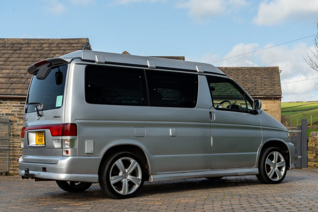 A 2006 Mazda Bongo Friendee silver van is parked on a stone-paved driveway. The van has tinted windows and a roof extension, featuring sleek alloy wheels. In the background, there are stone buildings and a cloudy blue sky. The license plate is visible at the rear of the van.