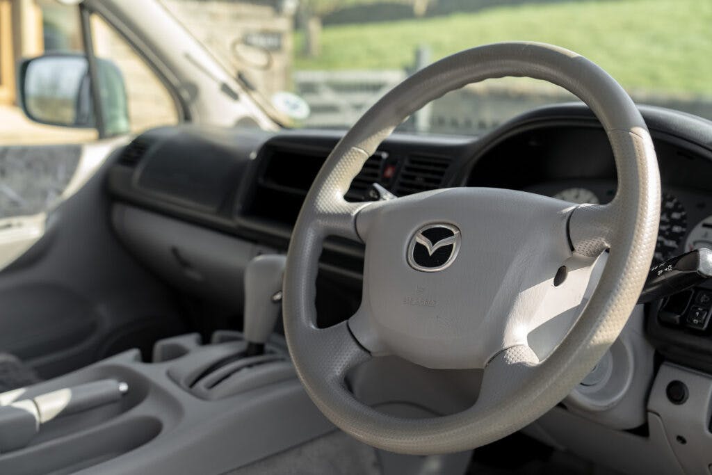 Interior view of a 2006 Mazda Bongo Friendee featuring a gray leather steering wheel with the Mazda logo. The dashboard is visible with an array of controls, air vents, and an instrument cluster. The car's interior appears clean and well-maintained.