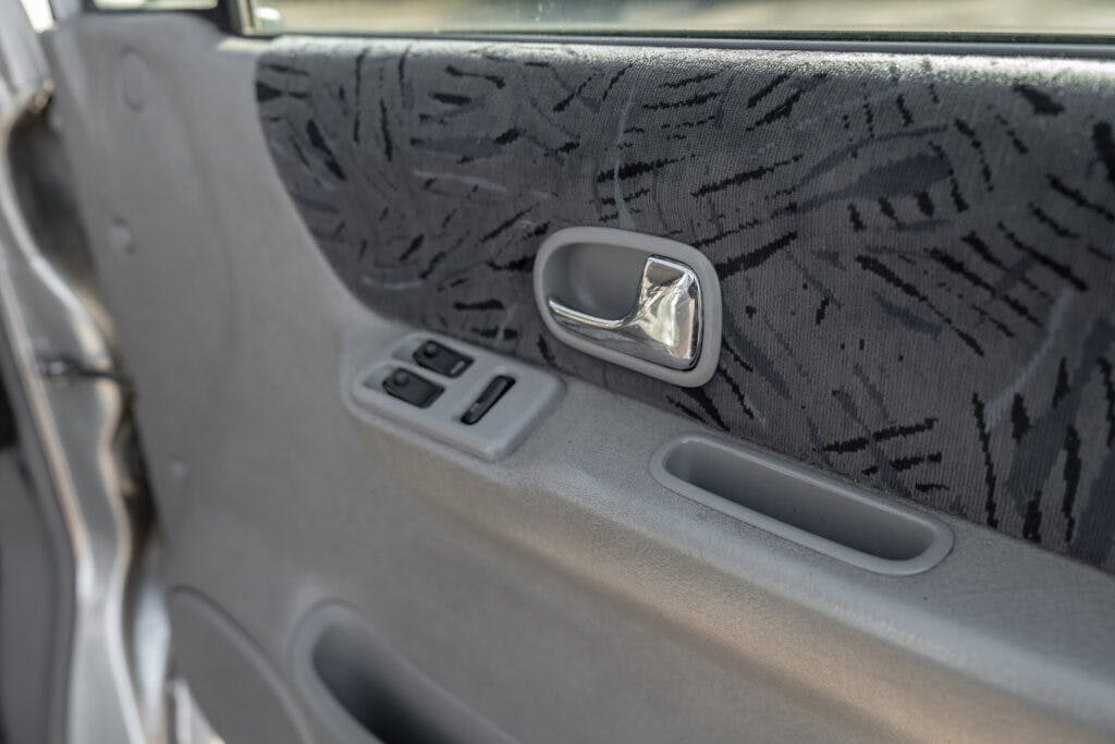Close-up view of a 2006 Mazda Bongo Friendee car door interior, featuring a silver door handle, power window control buttons, and textured dark fabric with a patterned design. The light gray door panel also includes a small compartment below the controls.