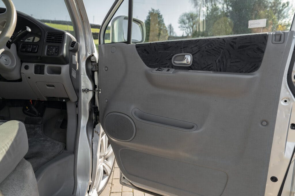 The interior of a 2006 Mazda Bongo Friendee is shown with the driver's side door open. The door panel features a handle, a speaker, and some patterned fabric. The dashboard, steering wheel, and pedals are visible, and the exterior scene includes some greenery.