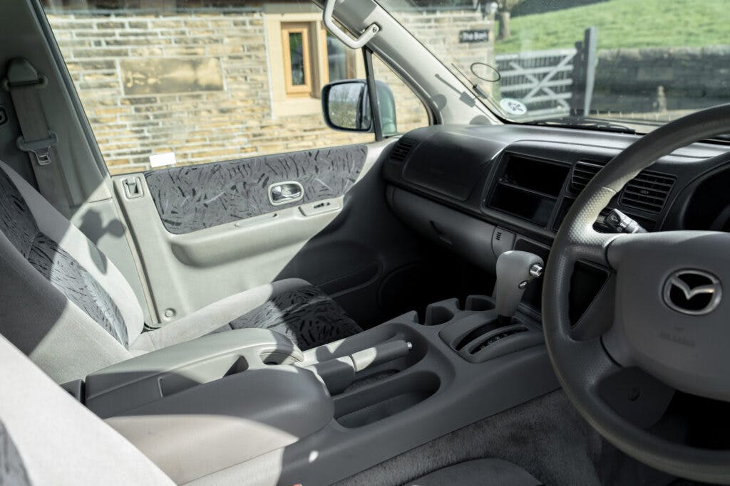 The interior of a 2006 Mazda Bongo Friendee shows the steering wheel, dashboard, center console, and two front seats. The sun shines through the window, illuminating the grey tones of the seats and door panel. A building wall and a wooden gate are visible outside the window.