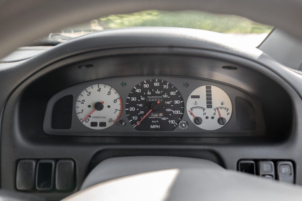 A close-up view of a 2006 Mazda Bongo Friendee dashboard shows the speedometer, odometer, tachometer, fuel gauge, and temperature gauge. The speedometer reads 0 MPH and the odometer displays 190,028 miles. The fuel gauge indicates about three-quarters full.