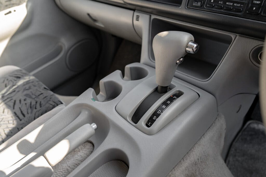 The image shows the interior of a 2006 Mazda Bongo Friendee, focusing on the center console. It includes an automatic gear shift lever, cup holders, and various buttons on the dashboard. The seats and surrounding surfaces are in shades of grey. The car appears clean and well-maintained.