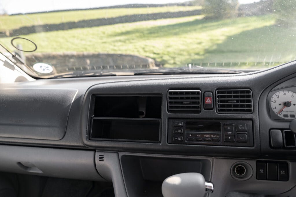 The interior of a 2006 Mazda Bongo Friendee dashboard shows an empty slot where an audio or video unit might be installed. The dashboard features air vents, climate control buttons, hazard light button, and a portion of the steering wheel. A grassy field is visible through the windshield.