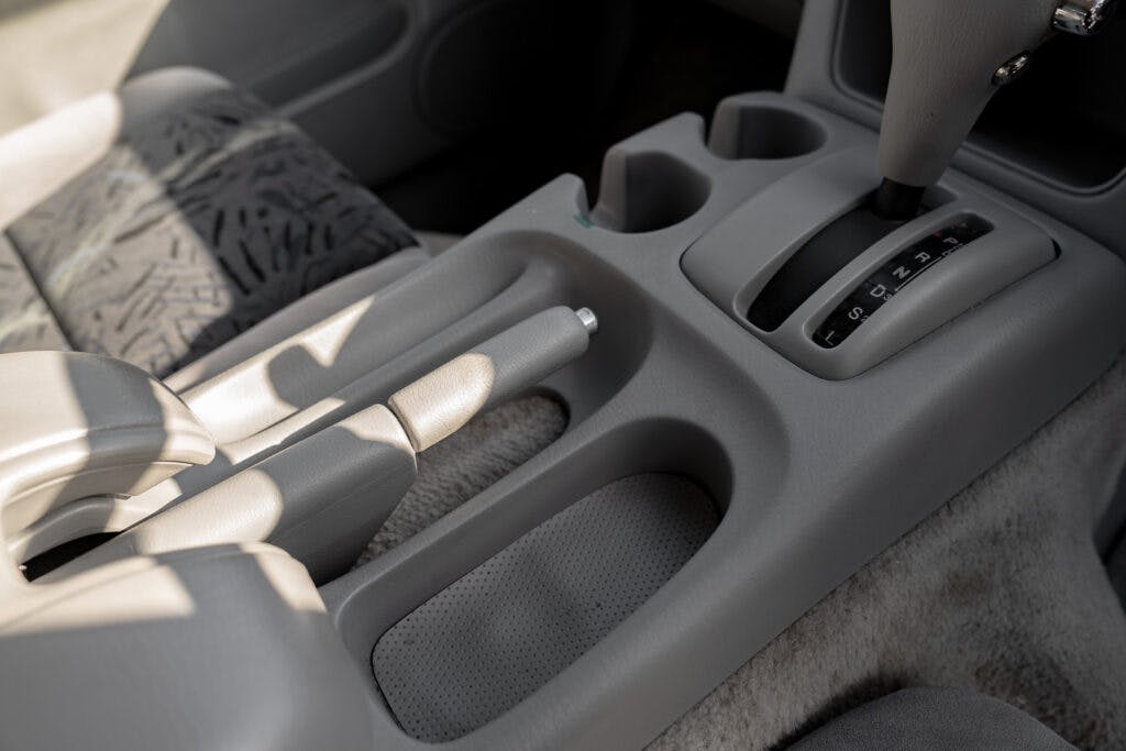 The image shows the center console of a 2006 Mazda Bongo Friendee's interior. Visible are the gear shift lever, cup holders, and part of the parking brake lever. The upholstery features a patterned fabric, and the overall color scheme is gray.