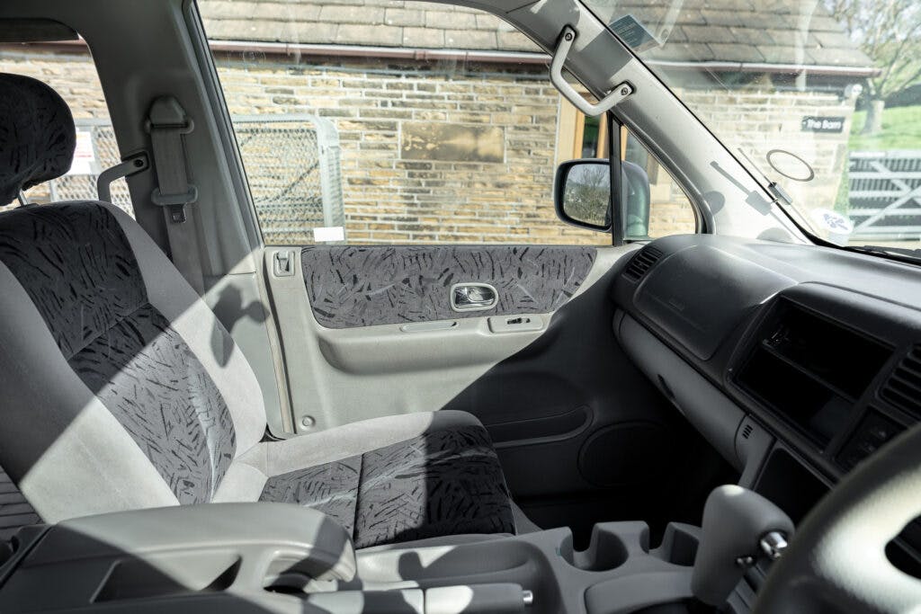Interior of a 2006 Mazda Bongo Friendee's cabin showing the front passenger seat and driver's seat. The cabin features dark patterned upholstery, manual window controls, a steering wheel, dashboard with built-in storage compartments, and cup holders. The window shows a brick wall outside.