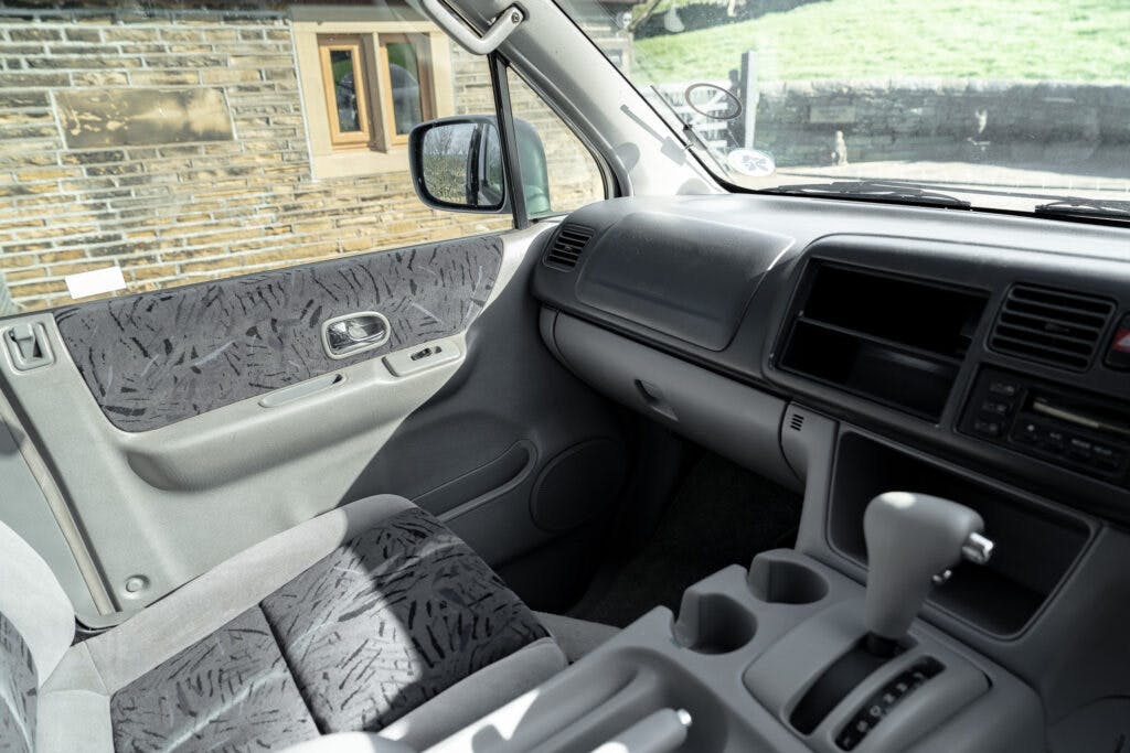 Interior of a 2006 Mazda Bongo Friendee with grey fabric seats and panel trim, featuring a dashboard with a steering wheel on the right side. The door panel shows patterned upholstery. Sunlight streams through the windows, illuminating the interior while a brick building is visible outside.