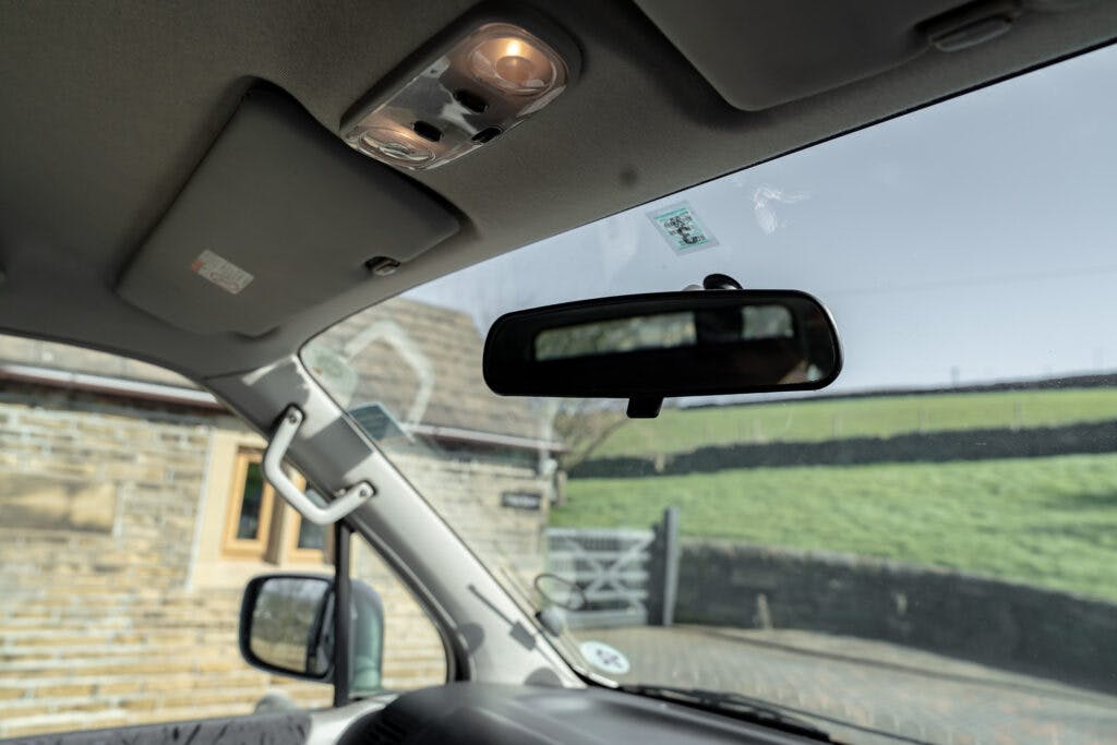 Interior view of a 2006 Mazda Bongo Friendee showing the windshield, rearview mirror, and part of the dashboard. A brick building with a window and a field with a stone wall are visible outside through the windshield. The car's ceiling light is on.