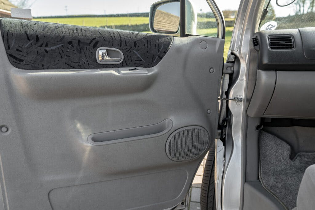 The interior of a 2006 Mazda Bongo Friendee is shown with a focus on the driver's side door. The door is gray with a textured panel, a silver handle, an integrated speaker, and a small storage compartment. The bottom part of the dashboard and a portion of the car's exterior are also visible.