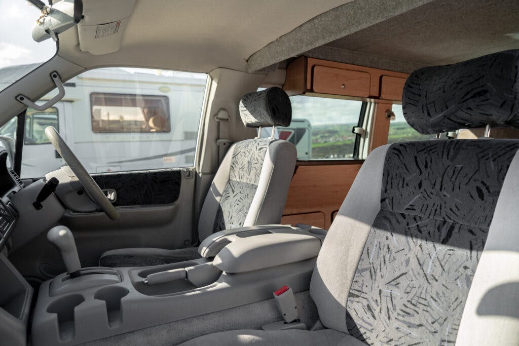 The image shows the interior of a 2006 Mazda Bongo Friendee with two upholstered front seats, a center console with a cup holder, and a wooden cabinet on the side. The back window reveals a view of another camper van and some greenery in the background.