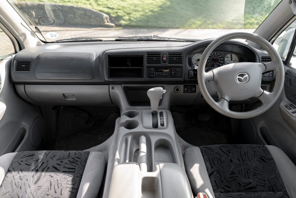 The image shows the interior of a 2006 Mazda Bongo Friendee, focusing on the dashboard, steering wheel, and front seats. The dashboard features controls, air vents, and storage compartments. The interior is light-colored with fabric seats displaying a pattern.