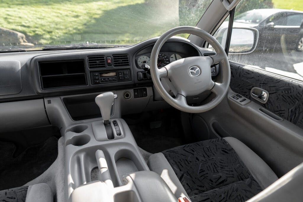 Interior view of a 2006 Mazda Bongo Friendee showing the driver's seat, steering wheel, dashboard, and center console. Features include a digital and analog display, air conditioning controls, a gear shift lever, and cup holders. The interior upholstery is patterned fabric.