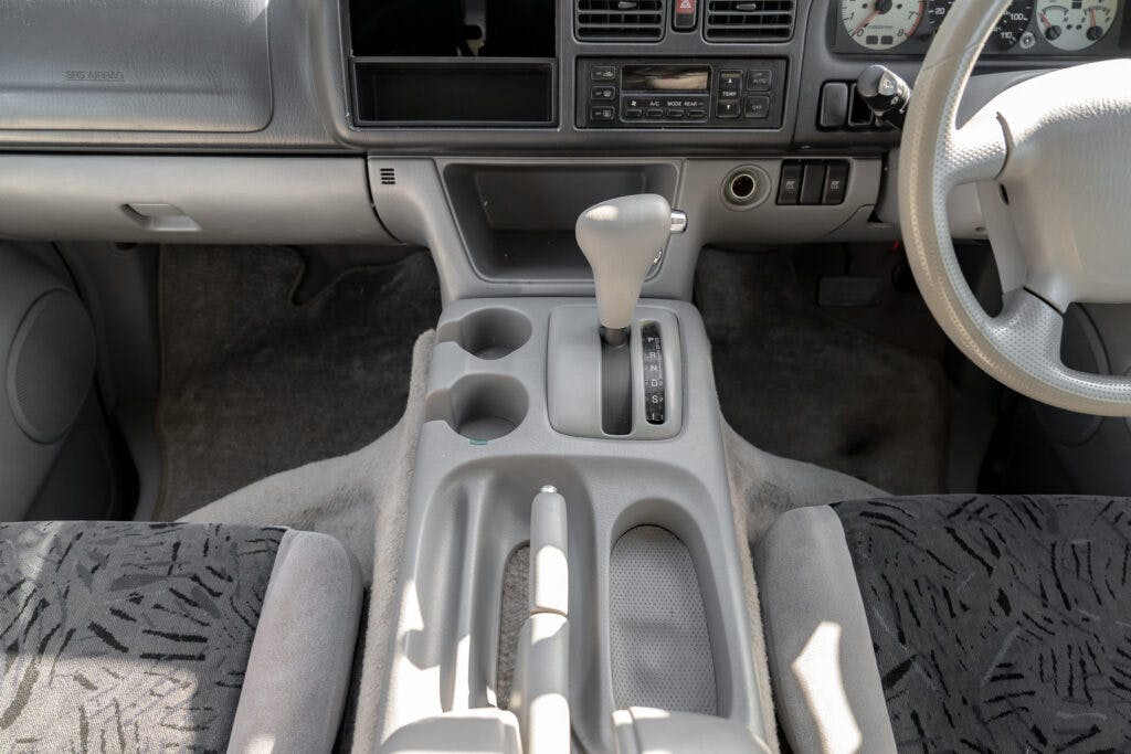 The image shows the interior of a 2006 Mazda Bongo Friendee, focusing on the center console and dashboard. The center console features a gear shift, cupholders, and storage compartments. The dashboard has various controls and a display screen. The seats have patterned covers.
