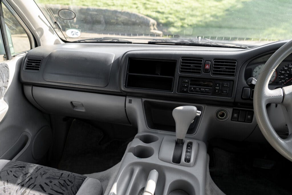 Interior view of a 2006 Mazda Bongo Friendee dashboard showing a steering wheel, gear shift, air vents, and a console with various controls and buttons. The seats have patterned upholstery, and sunlight is coming through the windshield.