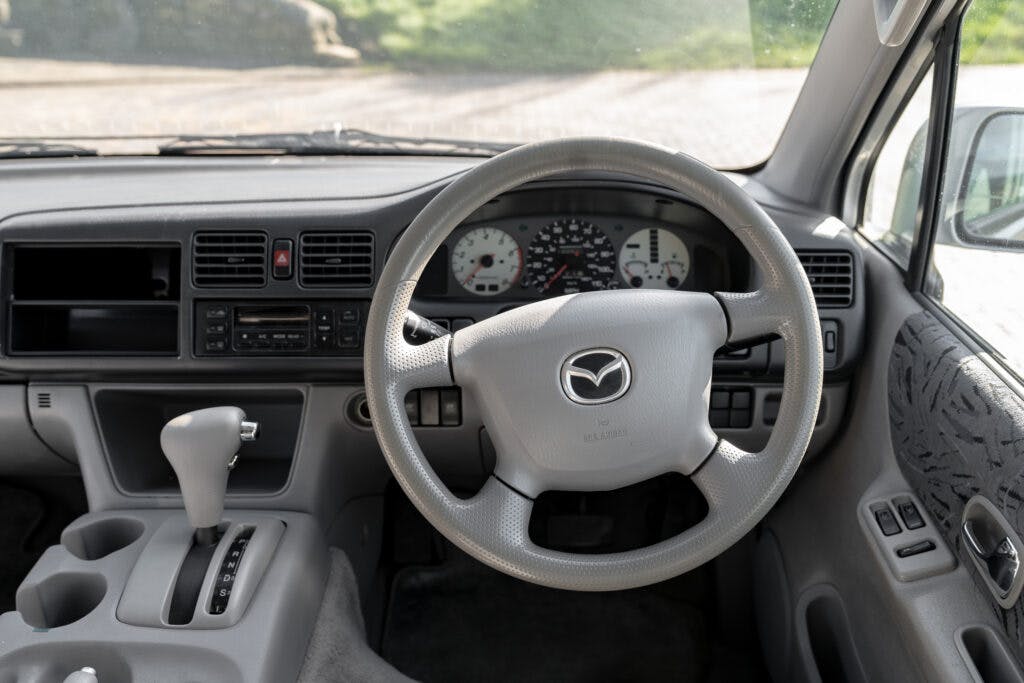 Interior view of a 2006 Mazda Bongo Friendee showing the driver's seat, dashboard, and steering wheel featuring the Mazda logo. The display panel includes a speedometer, tachometer, and fuel gauge. The gear shift is positioned on the console, and various controls are visible on the dash.