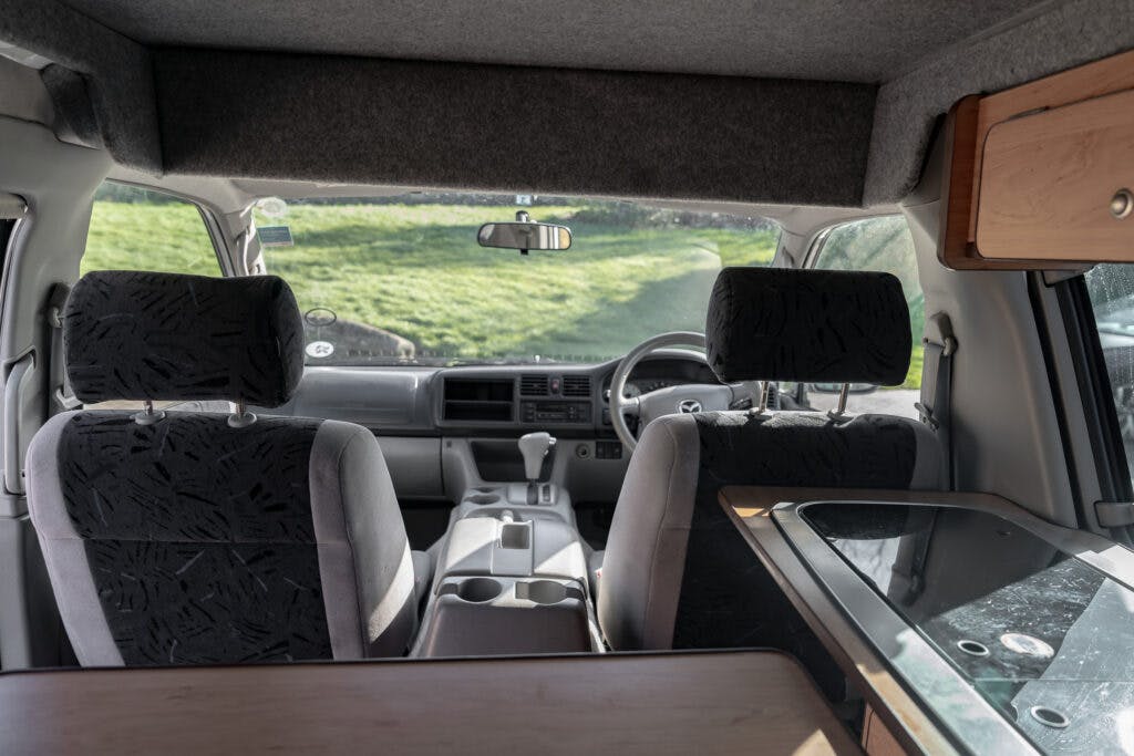 Interior view of a 2006 Mazda Bongo Friendee campervan showing the driver's and passenger's seats with patterned covers, the steering wheel, and the dashboard. The right side of the image includes a small wooden counter and a stove with a glass cover. Greenery is visible outside.