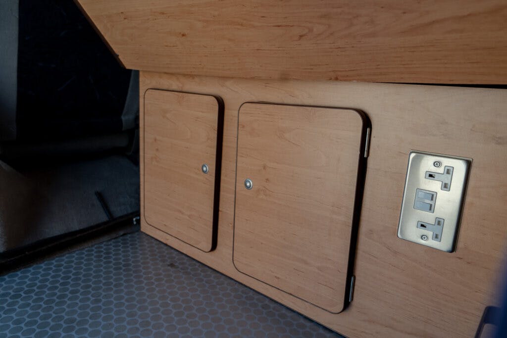 The image shows a section of a wooden interior with two closed rectangular compartments and a double electrical outlet with two switches, installed on the wooden surface. The floor, reminiscent of the interior's charm, has a hexagonal pattern that echoes the practicality seen in a 2006 Mazda Bongo Friendee.