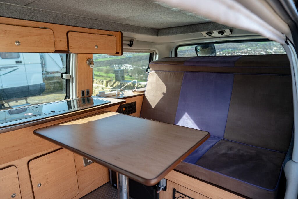 The 2006 Mazda Bongo Friendee camper van interior features a small wooden table attached to the kitchenette, complete with a sink and stove. Next to the table is a cushioned bench adorned with brown and blue upholstery. Cabinets and a window serve as the backdrop.