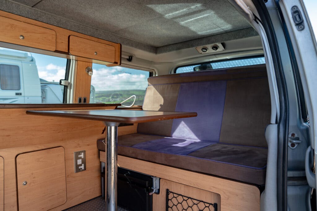 The image showcases the interior of a converted 2006 Mazda Bongo Friendee camper van with a small dining area. It features a wooden table attached to the floor and seating with blue and brown upholstery. Wooden cabinets adorn the walls, and a window provides ample natural light.