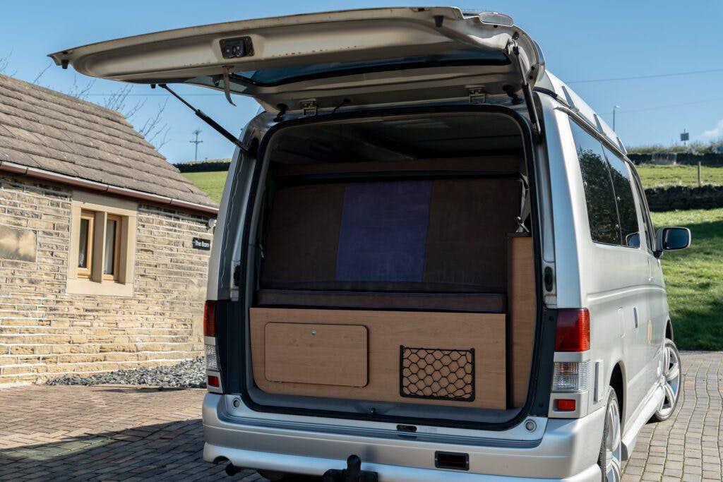 A silver 2006 Mazda Bongo Friendee with the rear door open reveals an organized interior setup, including a wooden storage unit with a mesh front compartment. The van is parked on a brick driveway near a stone building, with grass and an electric pole visible in the background.