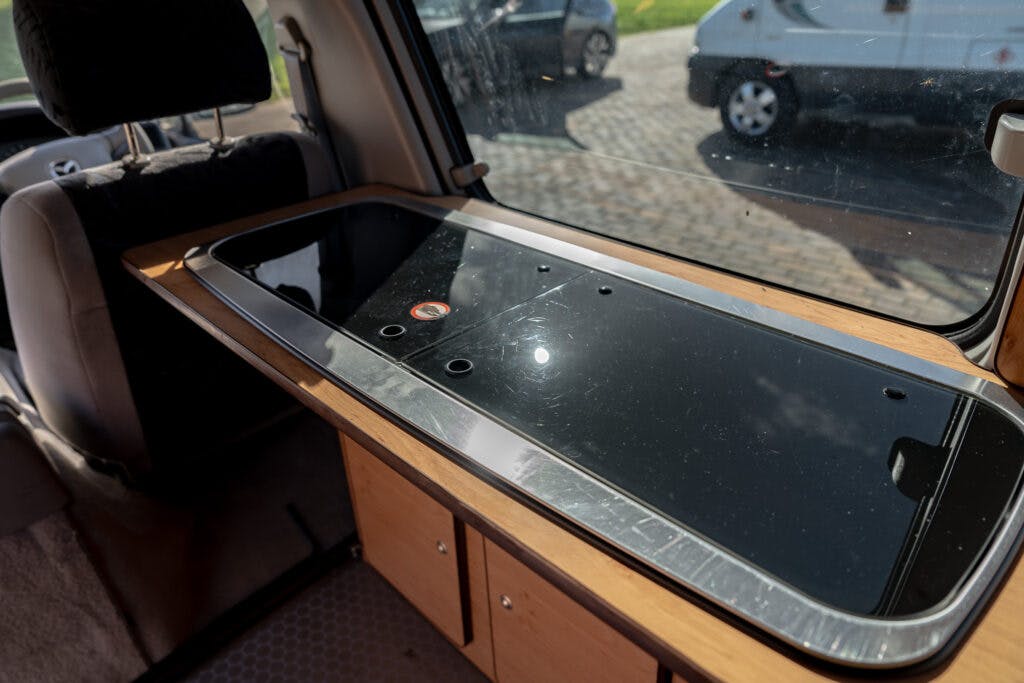 A photo shows the interior of a 2006 Mazda Bongo Friendee, featuring a sleek black countertop with several round holes along its length. Below the countertop, wooden cabinets and drawers provide ample storage. A window in the van reveals a parking area with some parked cars outside.