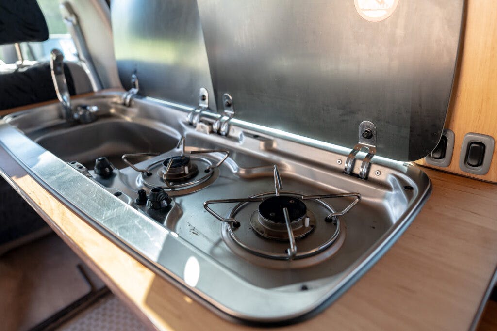 A close-up view of a built-in van kitchen setup in a 2006 Mazda Bongo Friendee, featuring a stainless steel sink on the left and a two-burner gas stove on the right. The cover for the stove is open, revealing the burners. The countertop is made of light-colored wood.