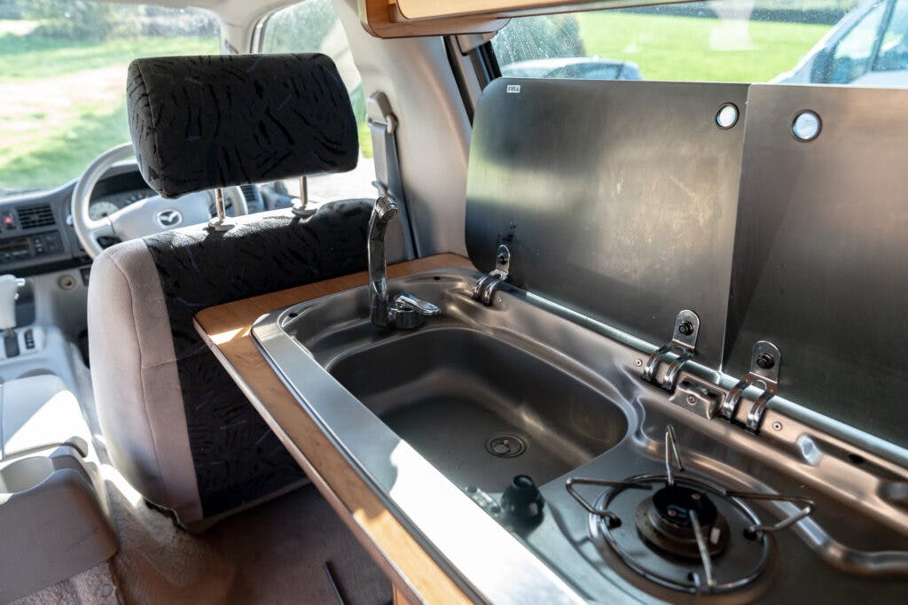 The image shows the inside of a 2006 Mazda Bongo Friendee, focusing on a compact kitchen setup. There is a stainless steel sink with a faucet and a single-burner stove. The kitchen area has hinged covers that can be folded down when not in use. The driver’s seat is visible in the background.
