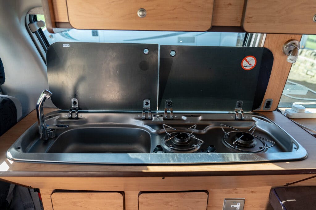 A compact kitchen setup inside a 2006 Mazda Bongo Friendee camper van features a sink on the left and two gas burners on the right. It has a metallic cover partially opened, wooden cabinetry, and a small window in the background. A "no smoking" sign is visible above the burners.