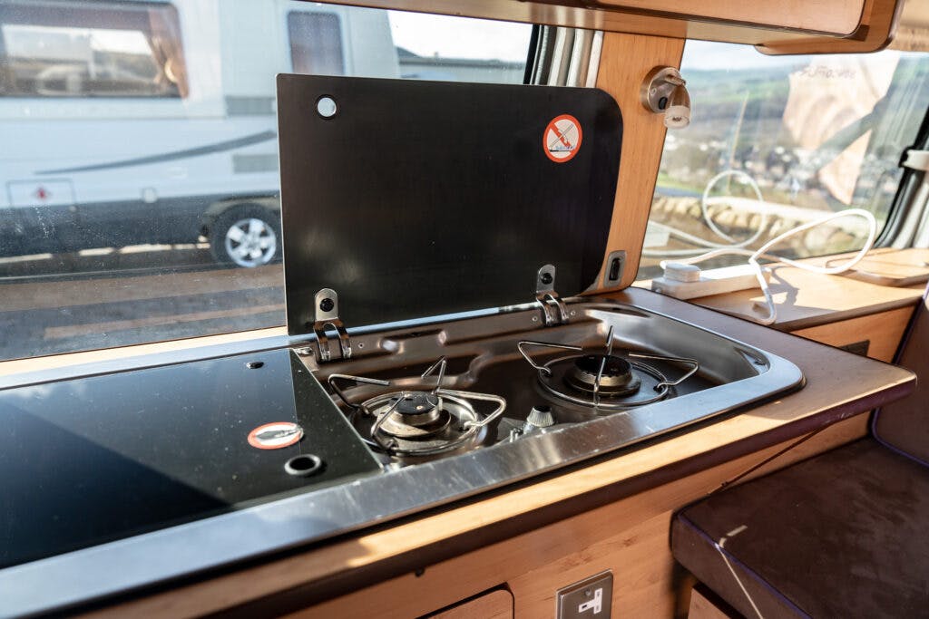 The image shows a compact kitchen area inside a 2006 Mazda Bongo Friendee camper van. It features a two-burner gas stove with a black lid that is propped open. A window in the background reveals another camper van and a landscape outside.