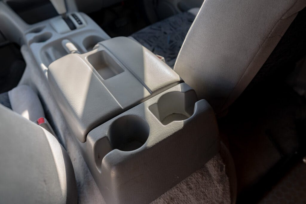 A close-up of a 2006 Mazda Bongo Friendee's center console between the front seats, showing two cup holders and a storage compartment. The console has a light gray color, and sunlight is casting shadows on it. The surrounding seats are partially visible.
