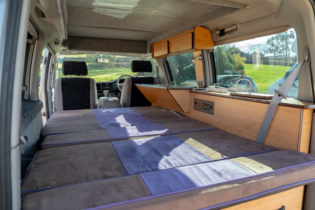 Interior of a 2006 Mazda Bongo Friendee converted into a camper with a folded-out bed covering the floor. Shelving and storage compartments are visible on one side. The front driver and passenger seats are partially visible in the background through a partition. Natural light streams in through multiple windows.