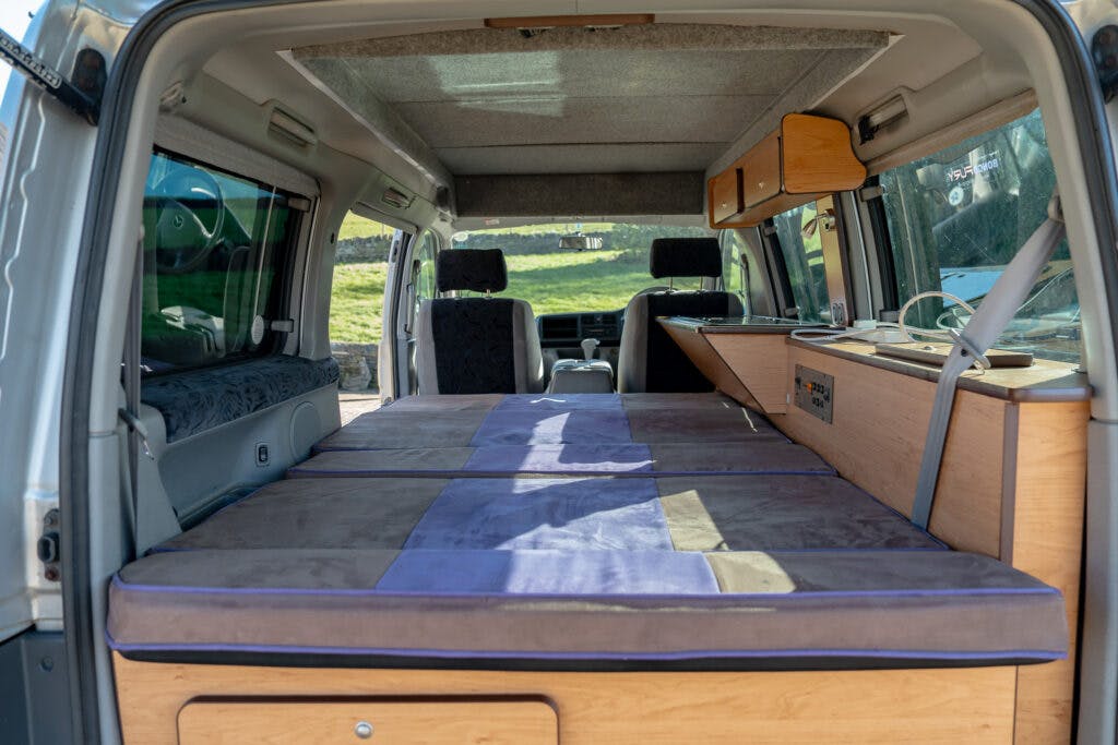The interior of a 2006 Mazda Bongo Friendee is shown, featuring a neatly made bed with cushions and pillows. The van also has wooden cabinets, storage compartments, and a window with a curtain. The driver and passenger seats are visible in the background.