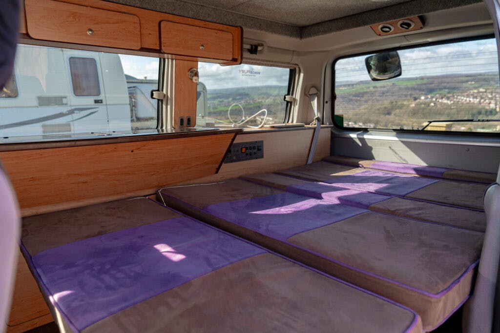 Interior of a 2006 Mazda Bongo Friendee with a fold-out bed covered in a purple and gray cushion. Wooden cabinets are mounted above the bed, and a window reveals another camper van and a landscape with hills and houses in the distance.