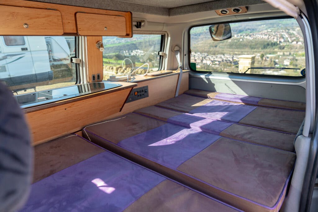 The interior of the 2006 Mazda Bongo Friendee showcases a converted sleeping area. The van features a flat bed with a patchwork of purple and beige cushions, overhead wooden cabinets, and a built-in power control panel. Sunlight streams through the rear and side windows.