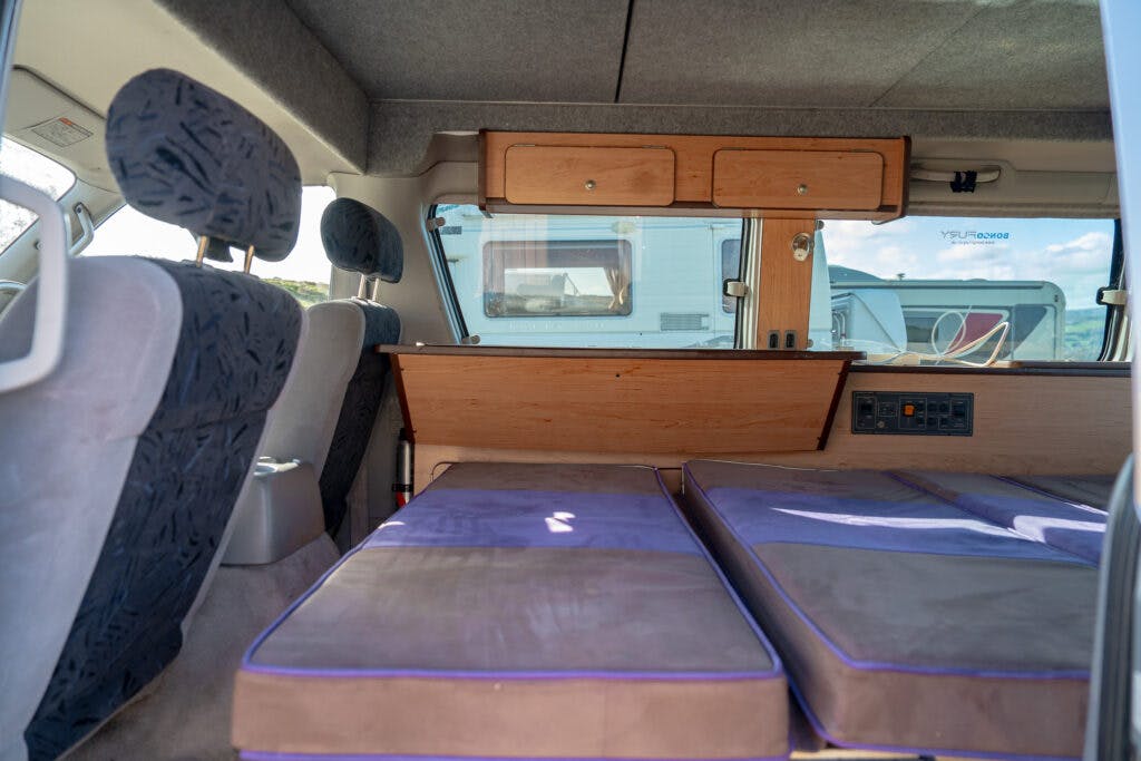 Interior of a 2006 Mazda Bongo Friendee with folded-down seats forming a bed. The arrangement includes two side-by-side cushioned sections covered in purple fabric. Overhead, there are two wooden storage cabinets attached to the vehicle’s ceiling.