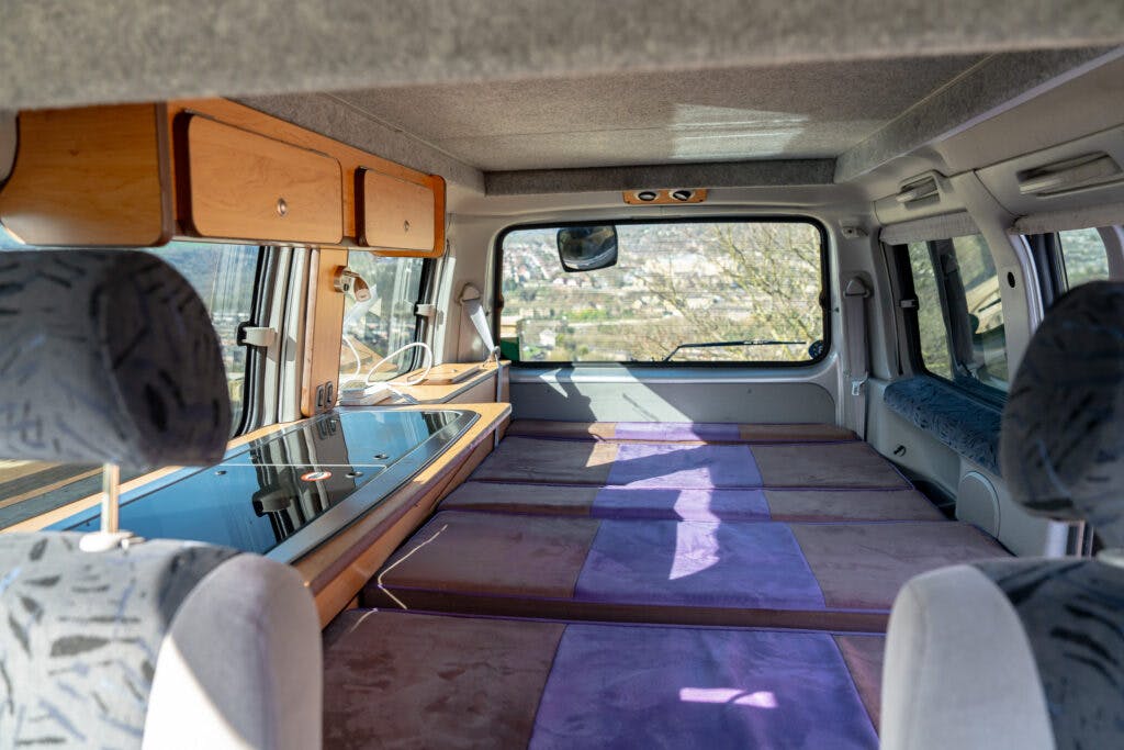 Interior view of a 2006 Mazda Bongo Friendee camper van showing a cozy sleeping area with purple pads arranged to form a bed. Wooden overhead storage cabinets are mounted on one side, and a window provides a view of the outdoors. The front driver and passenger seats are partially visible.