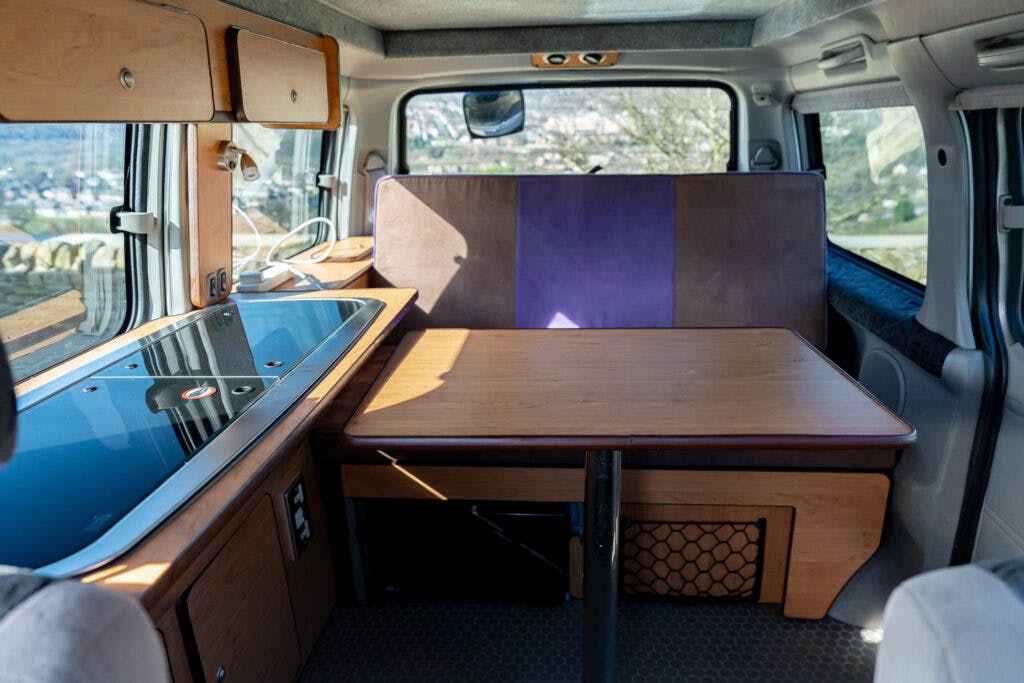Interior of a 2006 Mazda Bongo Friendee camper van showing a wooden dinette table and cushioned seating area, with windows providing natural light. The kitchenette area includes a stovetop, sink, and overhead storage compartments. The seats and table have a combination of brown and purple shades.