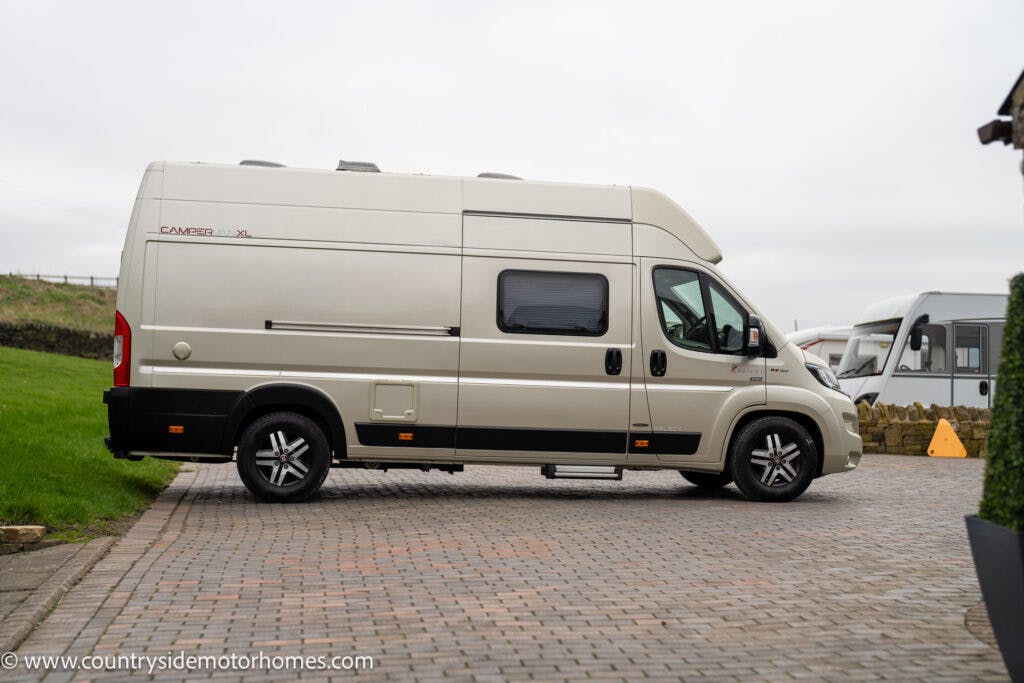 A beige 2020 Rapido Dreamer Select Campervan XL is parked on a paved driveway. The campervan features a high roof, large windows, and sleek black alloy wheels. In the background, part of another vehicle and a stone wall are visible under an overcast sky.