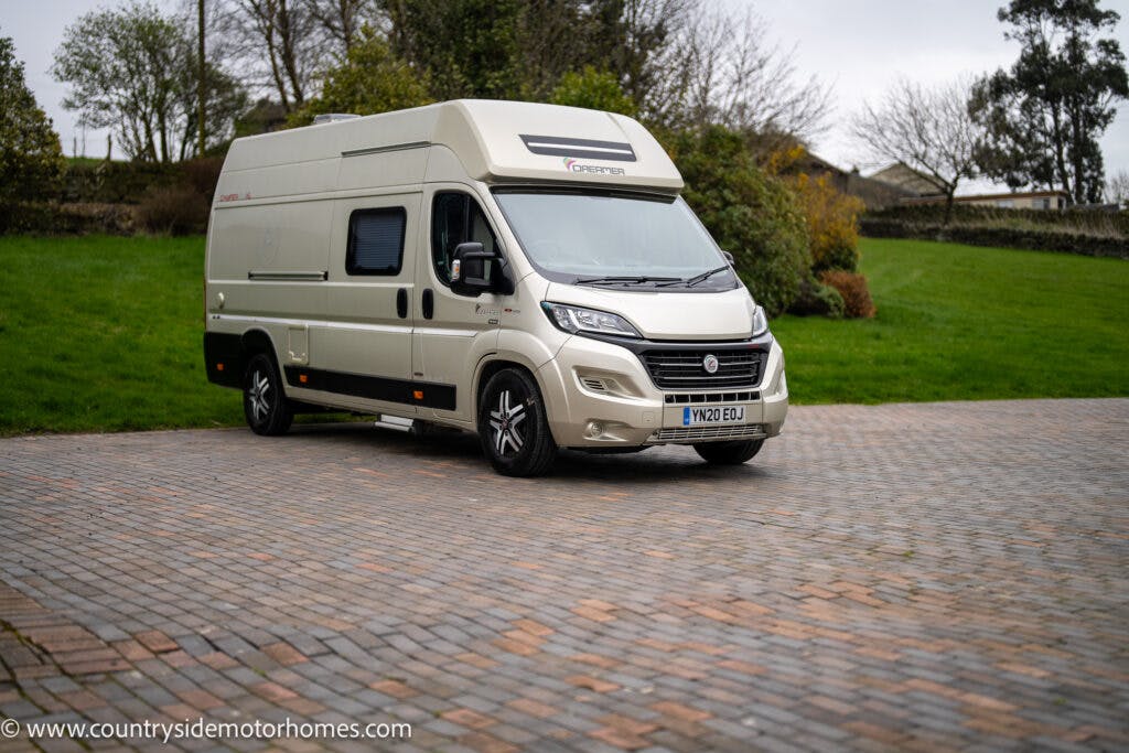 A white 2020 Rapido Dreamer Select Campervan XL with a high roof is parked on a paved driveway against a backdrop of green grass and trees. The vehicle has the registration plate VN20 EOU and a logo on the side. The overcast sky suggests it might be a cloudy day.