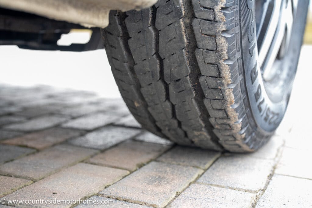 Close-up of a vehicle tire on a paved surface. The tire shows signs of wear with visible tread patterns, and there is some mud and dirt on the tire's surface. The image also captures part of the underside and axle of the 2020 Rapido Dreamer Select Campervan XL.