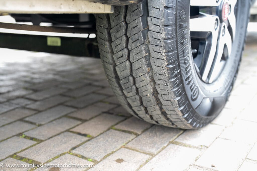 A close-up view of a vehicle tire on a brick-paved surface. The tire tread design is visible, and it is attached to the vehicle's wheel rim. The image shows part of the undercarriage of a 2020 Rapido Dreamer Select Campervan XL. The website "www.countrysidemotorhomes.com" is displayed.