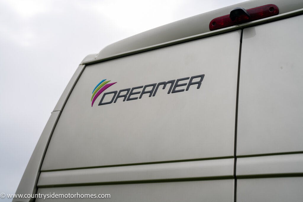 Close-up of the rear of a white 2020 Rapido Dreamer Select Campervan XL, adorned with a colorful "Dreamer" logo. The vehicle features a red tail light at the top center and panel lines. The background is slightly blurred, possibly a cloudy sky. Website URL visible at the bottom left corner.