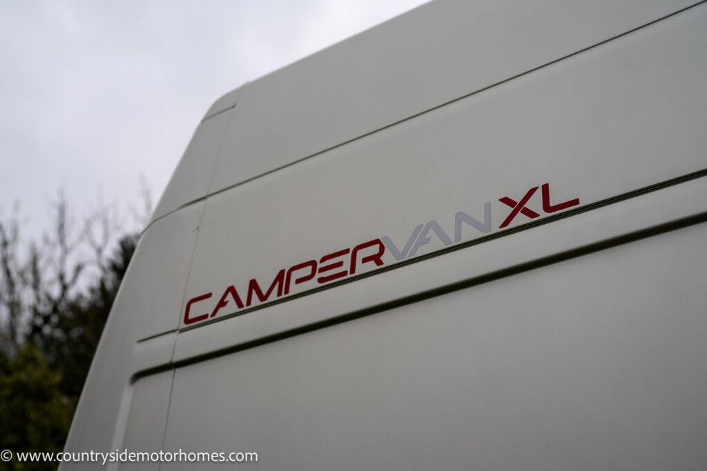 Close-up of the side of a white 2020 Rapido Dreamer Select Campervan XL displaying the model name "CAMPERVAN XL" in red and gray lettering. The background shows some out-of-focus trees and a cloudy sky. The URL "countrysidemotorhomes.com" is visible in the bottom left corner.