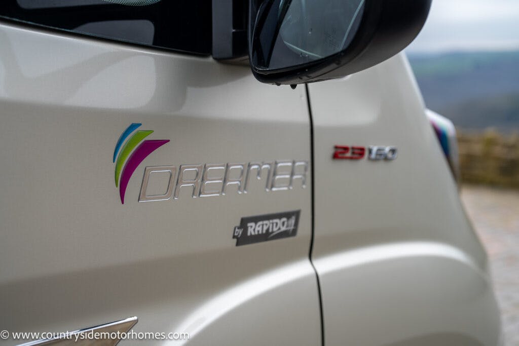 Close-up of a silver 2020 Rapido Dreamer Select Campervan XL showing the logo "Dreamer by Rapido" along with a multicolored emblem. The text "23 150" is also visible on the side. The reflection of a mirror and a blurred countryside landscape are seen in the background.
