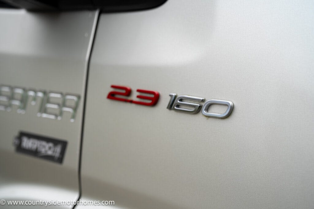 Close-up of a 2020 Rapido Dreamer Select Campervan XL's rear side, showcasing the "2.3 160" badge. The vehicle is silver, with "2.3" in red and "160" in silver. In the background, part of another badge and a blurred logo are visible.