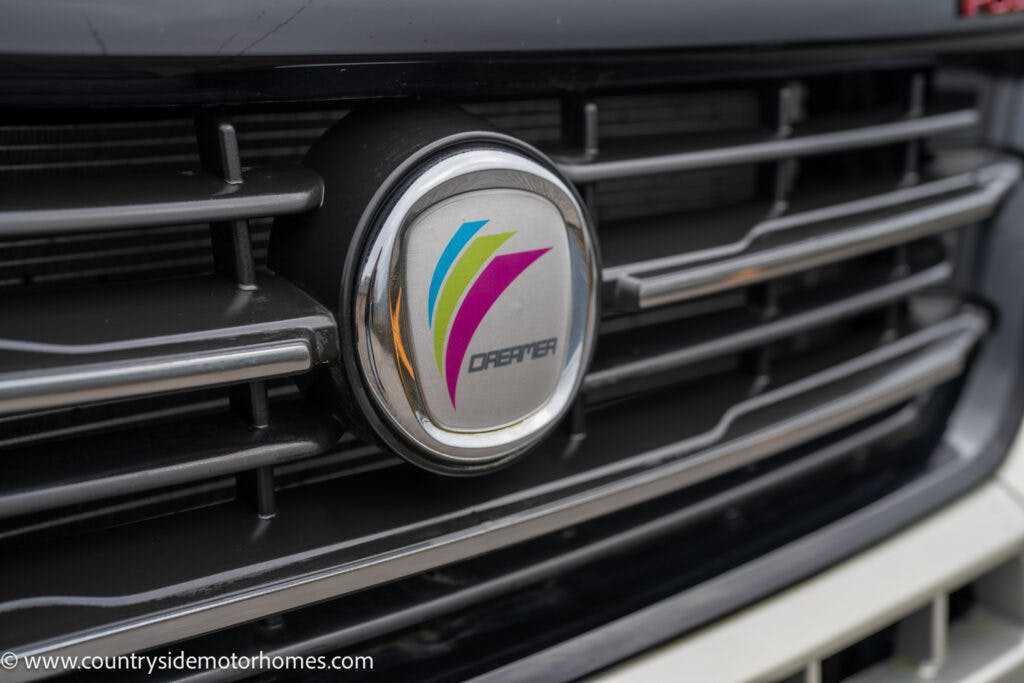 Close-up of a vehicle grill with a circular emblem bearing a multi-colored 'V' logo and the text "DREYER" in the center, reminiscent of the 2020 Rapido Dreamer Select Campervan XL. The web address www.countrysidemotorhomes.com is seen in the bottom left corner.