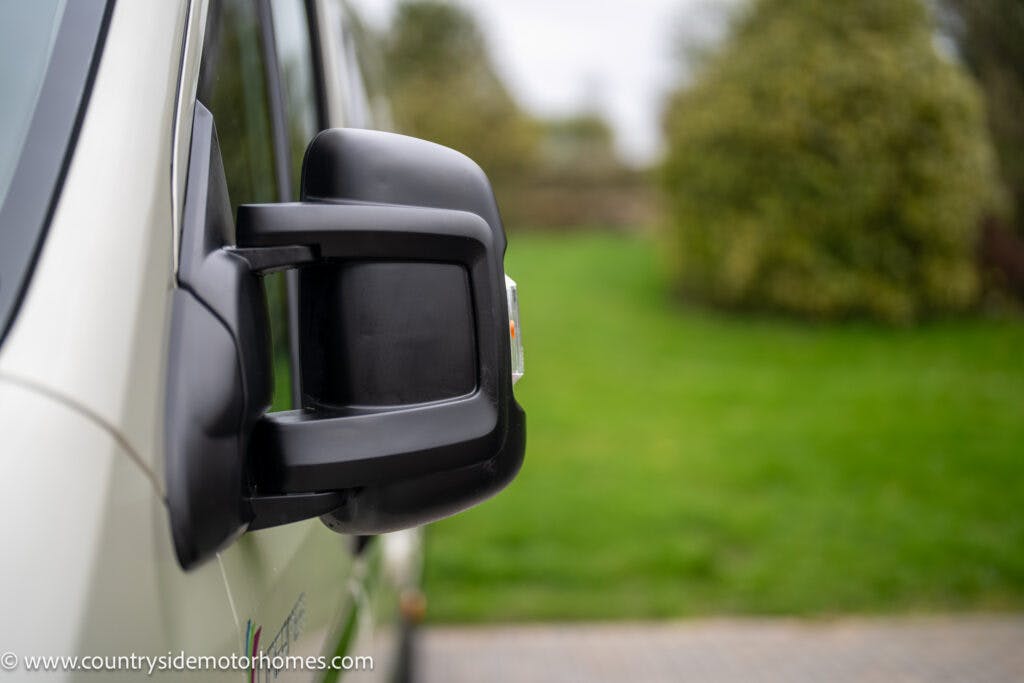 Close-up of a black side mirror on a white 2020 Rapido Dreamer Select Campervan XL with an amber indicator. The foreground shows the side of the vehicle, while the background features a blurred view of green grass and shrubbery. A URL, www.countrysidemotorhomes.com, is partly visible in the lower left.