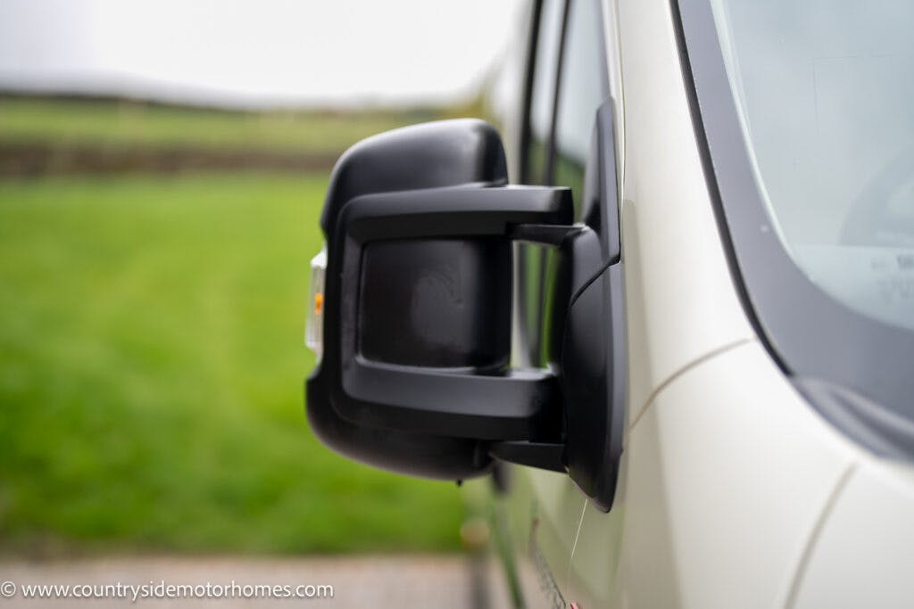 Close-up of a black side mirror attached to a white 2020 Rapido Dreamer Select Campervan XL. The background is slightly blurred, showing green grass and a distant fence. The image is taken during the daytime.